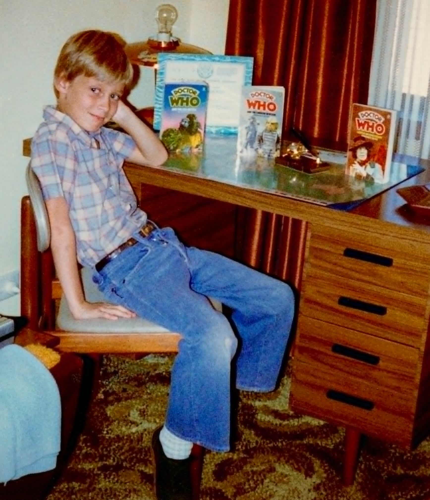 Peter is a little blond boy in a checked shirt and jeans, sitting at his desk. There's a framed certificate on his desk, and a wrapped present. There are also three novelisations: The Sea Devils, The Android Invasion, and The Invisible Enemy.