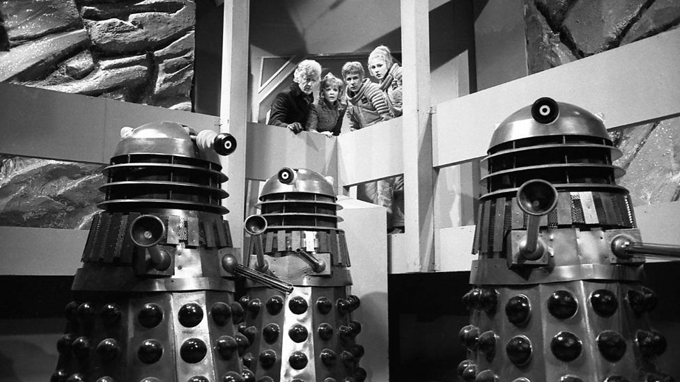 The Doctor, Jo, Codal and Rebec are standing on the walkway looking down at an army of three Daleks on the floor below.