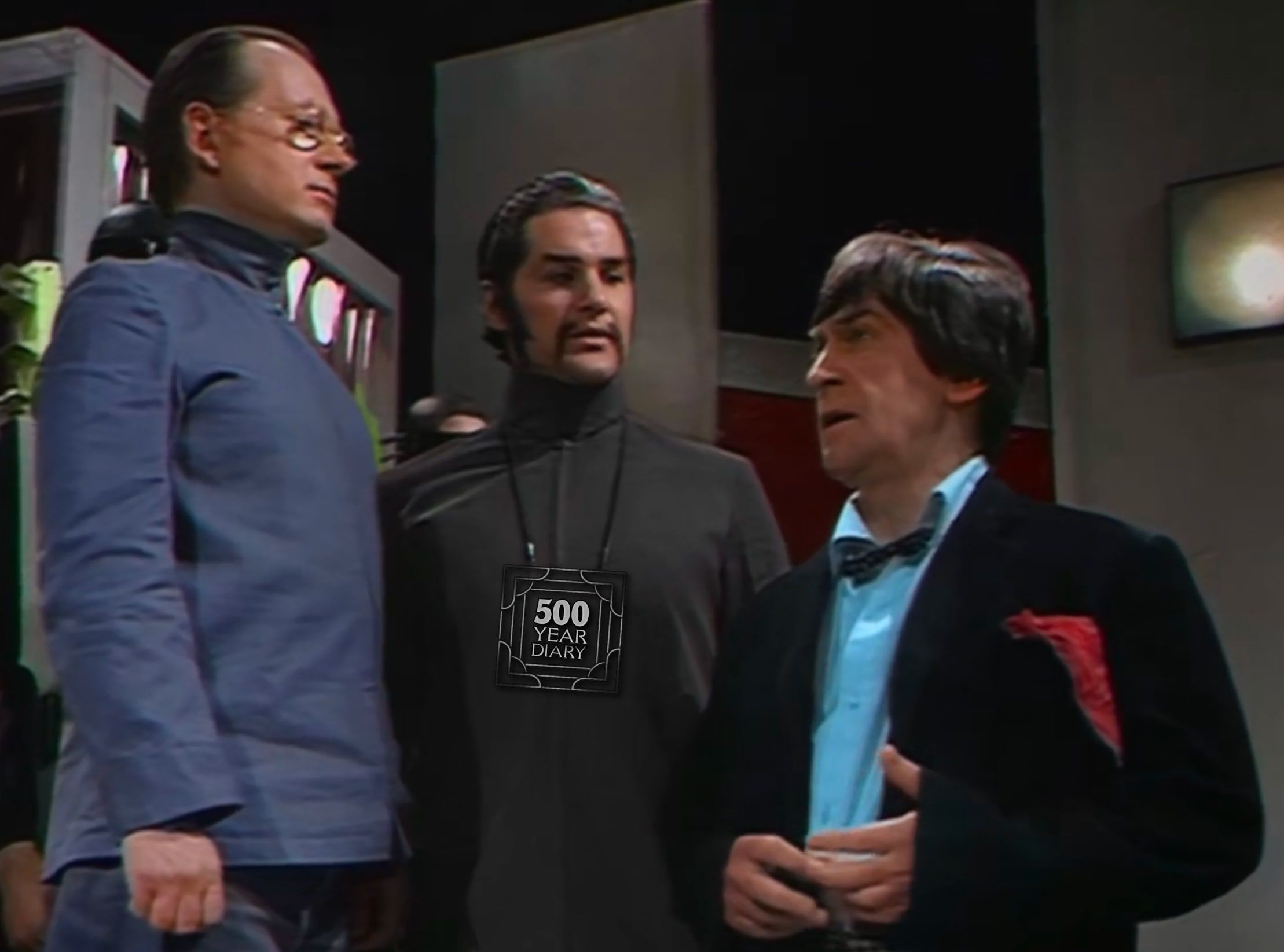 The Security Chief, the War Chief and the Doctor are standing and talking in the War Chief's headquarters. The War Chief is wearing a stylish pendant in the shape of the 500 Year Diary logo.