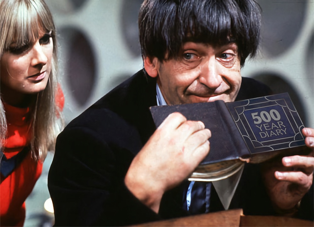 Patrick Troughton's Doctor is in the TARDIS, leaning on a wooden chest.  In his hands is a little book with the logo of the 500 Year Diary podcast  on the front. Polly is looking at it over his shoulder.